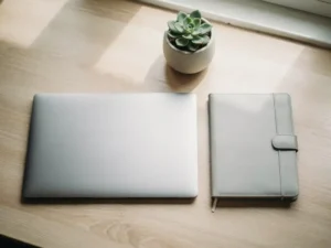 Laptop en leren agenda naast elkaar op een licht eikenhouten bureau, met natuurlijk daglicht en een kleine vetplant.