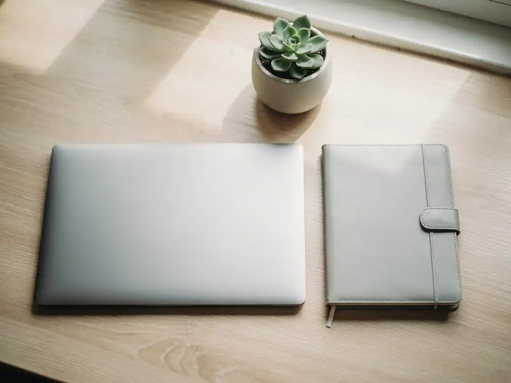 Laptop en leren agenda naast elkaar op een licht eikenhouten bureau, met natuurlijk daglicht en een kleine vetplant.